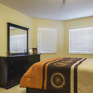A cozy bedroom with a dresser and mirror, featuring large windows with blinds.