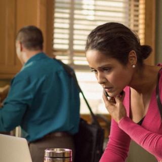A woman on a phone call in a kitchen while using a laptop, with a man in the background.