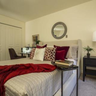 A cozy bedroom with elegant decor, featuring a bed adorned with red and white pillows, a side table with books, and a round mirror on the wall.
