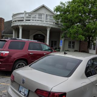 A view of a building with a balcony, surrounded by parked cars and trees under a cloudy sky.