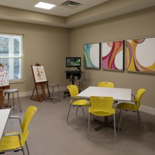 A bright and inviting art room featuring tables, chairs, easels, and colorful paintings.