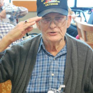 An elderly man wearing a veteran hat saluting indoors.