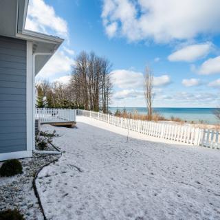 A snowy beach view with a house and fence near a lake under a partly cloudy sky.