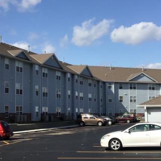 A residential apartment building with a parking lot in front under a blue sky with clouds.