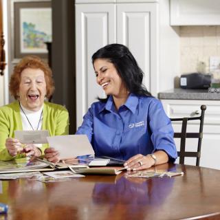 A caregiver and an elderly woman looking at photos together at a kitchen table, smiling and enjoying their conversation.