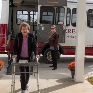 An elderly woman using a walker disembarks from a bus with a caregiver nearby, surrounded by autumn flowers.