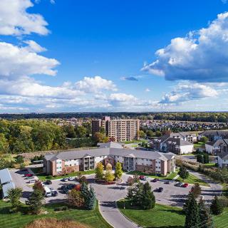 Aerial view of a suburban neighborhood with buildings, surrounded by trees under a cloudy blue sky.