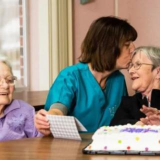 A caregiver shares a tender moment with elderly women at a celebration, accompanied by a cake on the table.