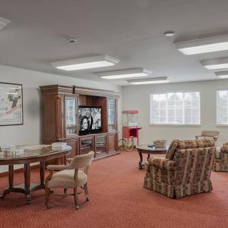 A cozy living room with chairs and a television, featuring a carpeted floor and large windows.