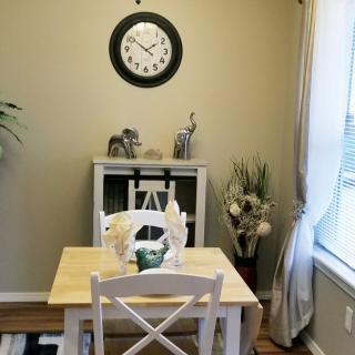 A cozy dining area with a small table and chairs, adorned with decorative items and plants, featuring a wall clock.