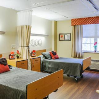 A hospital room with two beds, separated by a privacy curtain, featuring decorative pillows and a large window.