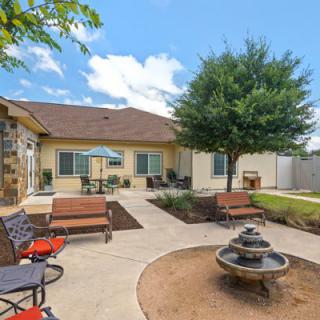 A beautiful backyard with a patio, seating area, and a small fountain surrounded by greenery on a sunny day.