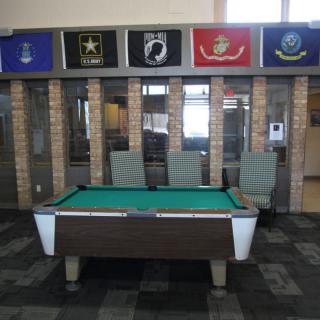 A pool table is set in front of a brick wall adorned with various military flags.
