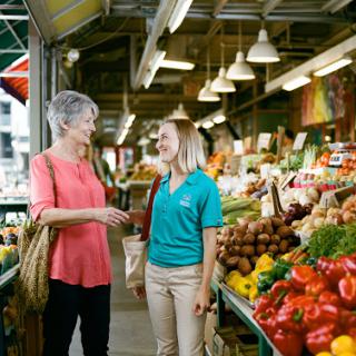 Two women engage in conversation at a vibrant market, surrounded by fresh fruits and vegetables.