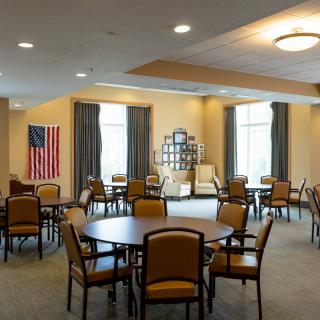 A well-lit dining room with multiple round tables and chairs, featuring an American flag on the wall.