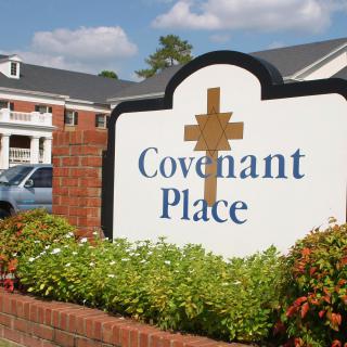 A sign for Covenant Place with a cross, situated in front of a building and surrounded by flowers.