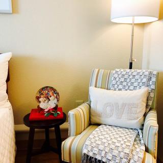 A cozy bedroom corner featuring a striped chair with a pillow and blanket, accompanied by a side table holding decorative items and illuminated by a standing lamp.