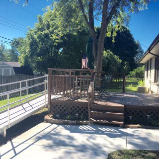 A sunny backyard scene with a wooden deck, accessible ramp, and lush green trees.