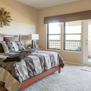 A well-lit bedroom featuring a neatly made bed with patterned bedding, large windows, and stylish decor.
