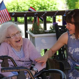 An elderly woman in a wheelchair shares a joyful conversation with a younger woman outdoors, surrounded by decorations and flags.
