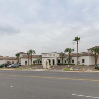 A building with palm trees lining the entrance, set against a cloudy sky and adjacent to a road.