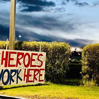 A sign reading 'Heroes Work Here' is displayed outdoors near bushes during the evening.