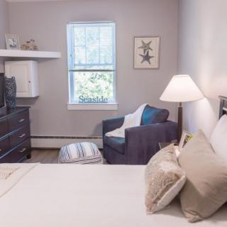 A cozy bedroom with a neatly made bed, an armchair by the window, and soft lighting from a bedside lamp.