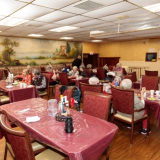 A busy restaurant interior with patrons dining at tables covered in maroon tablecloths.