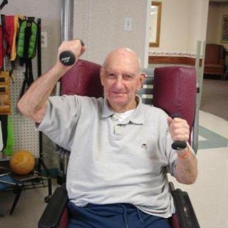 An elderly man exercising with dumbbells while seated in a chair.