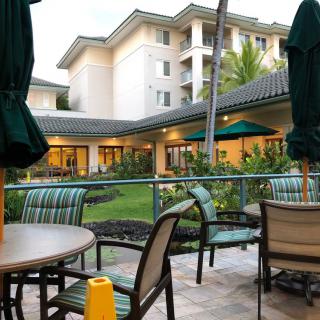 Outdoor patio area with tables, chairs, and umbrellas in a landscaped setting near a multi-story building.