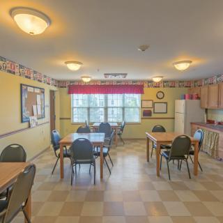 A cozy dining room with several tables and chairs, featuring a small kitchen area and large windows letting in natural light.
