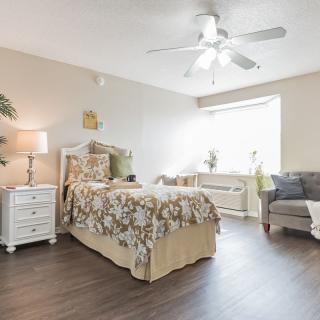 A cozy and well-lit bedroom with a floral-patterned bedspread, a bedside table with a lamp, and a comfortable chair by the window.