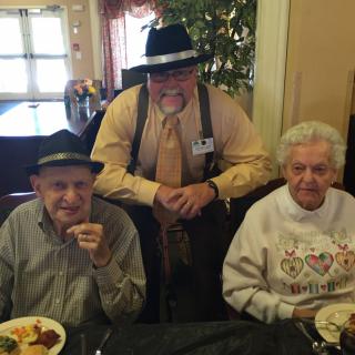 A group photo of three people at a dining table, two elderly individuals seated and one person standing behind them, all wearing hats.