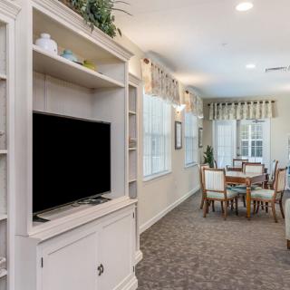 A cozy living and dining area featuring a television on a built-in shelving unit, with carpeted flooring and elegant drapes.