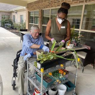 A healthcare worker assists a senior citizen in a wheelchair with gardening activities outdoors.