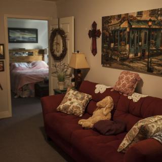 A cozy living room with a maroon sofa adorned with patterned pillows, artwork on the wall, and a view into an adjacent bedroom.