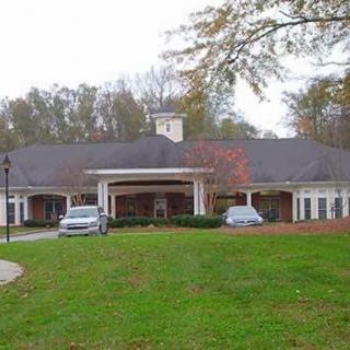 A large residential building with a driveway and parked cars, surrounded by trees and a lawn.