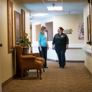 Two people walking and conversing in a hallway with artwork on the walls and furniture along the side.