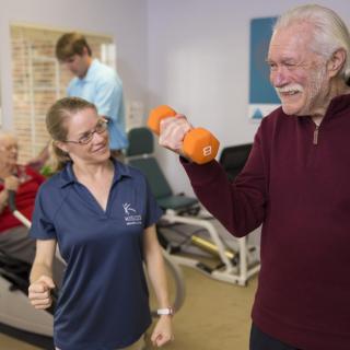 An elderly man lifting a dumbbell with the guidance of an instructor in a physical therapy setting.