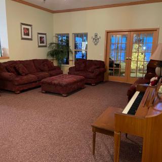 A cozy living room featuring a set of maroon sofas, a wooden piano, and large windows allowing natural light.