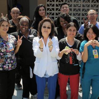 A diverse group of people posing together outdoors, showing positive gestures and smiling.