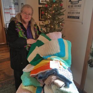 A woman standing indoors holding a collection of colorful crocheted blankets, with a tree in the background.