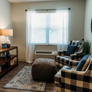 A cozy living room with plaid armchair and ottoman, illuminated by natural light from a large window.