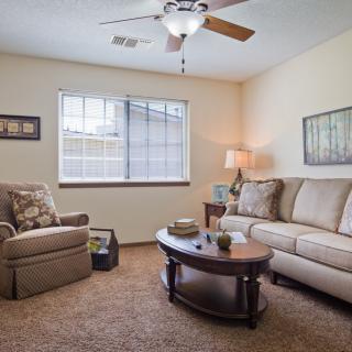 A cozy living room with a comfortable sofa, armchair, and coffee table, illuminated by a lamp and natural light from the window.