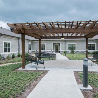 A landscaped outdoor area featuring a pergola and seating, leading to a building entrance.