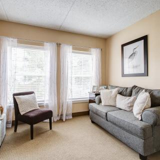 A cozy living room with a gray sofa, cushions, and a chair, illuminated by natural light through two large windows.