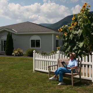 A woman reads on a bench in front of a house with sunflowers and a white picket fence.