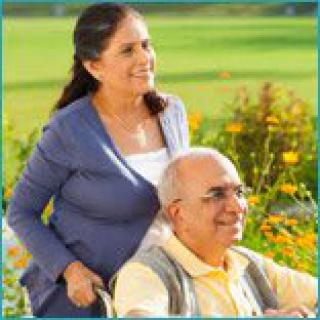 An elderly man in a wheelchair is being assisted by a woman in a sunny garden.