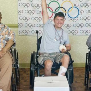 A person in a wheelchair celebrates with an Olympic-themed background and a medal.
