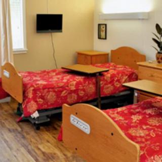 A well-lit hospital room featuring two twin beds with red floral covers, wooden furniture, and a wall-mounted TV.